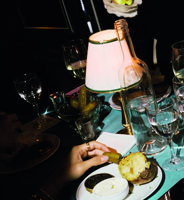 A candlelit table under a pleated lamp: glasses, caviar and a hand reaching for the plate.