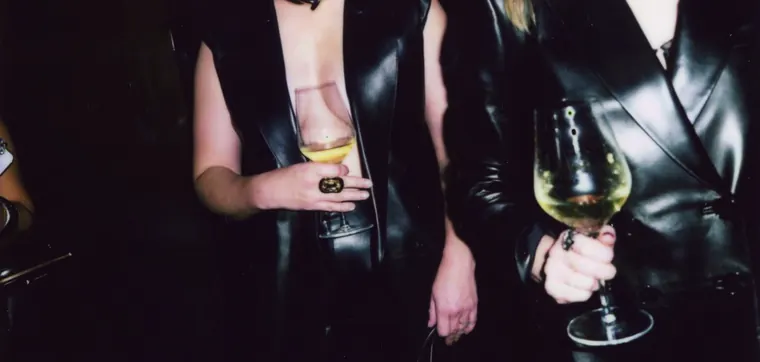 Instant photograph: two guests in black leather holding glasses of white wine.