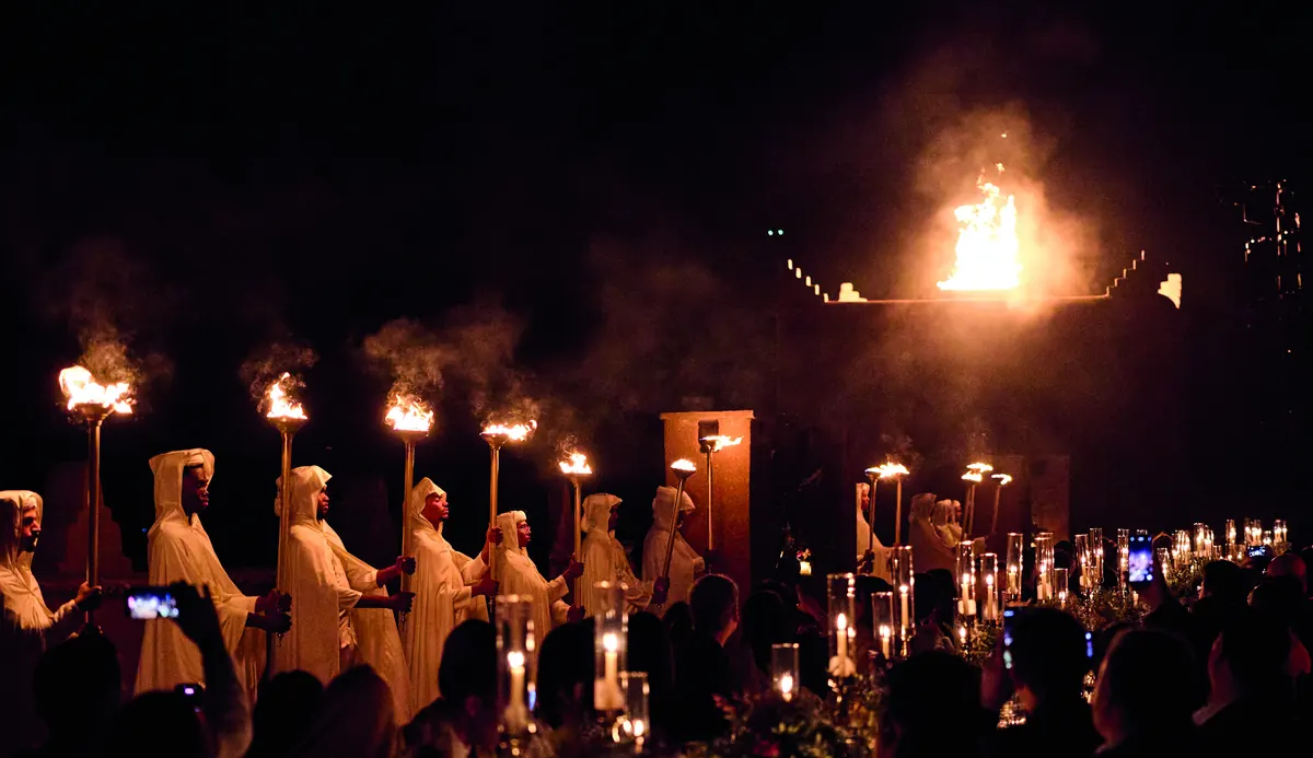 Hooded figures holding flaming torches above a candlelit dinner, at night.