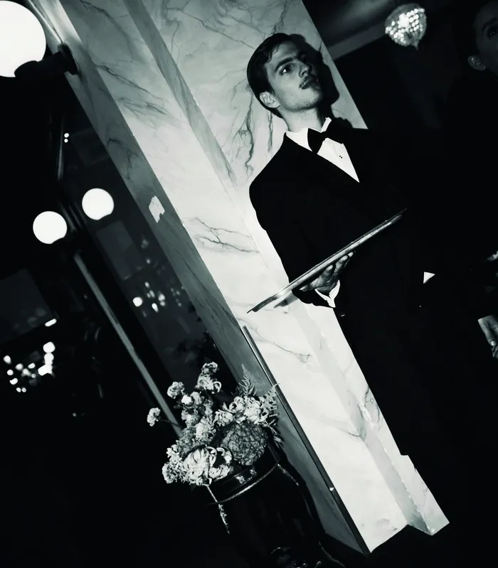 Black and white: a waiter in a tuxedo holding a silver tray beside a marble column.